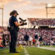A camera operator on a stadium sideline at golden hour using a lightweight camera rig with a compact wireless transmission unit.