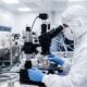 Photorealistic image of a cleanroom technician inspecting a spool of ultra-fine wire under a microscope station during medical coil manufacturing.