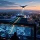 Illustration of a drone lifting off from a rooftop dock at dawn while operators in a command center watch live aerial video.